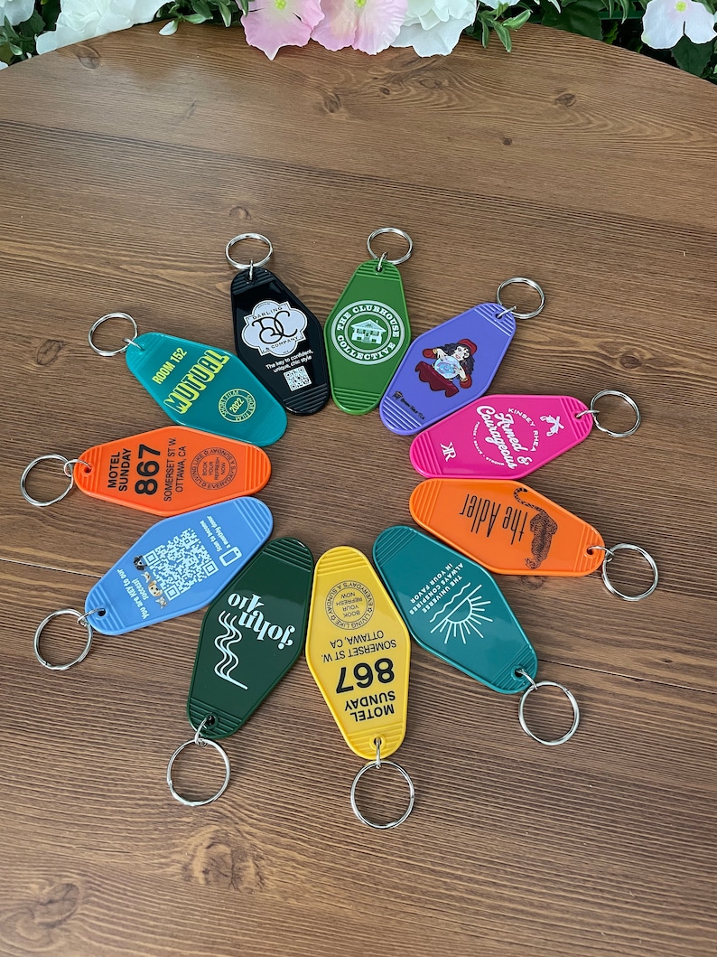 May include: A collection of ten colorful keychains with various designs. The keychains are shaped like motel keys and feature different text and graphics, including the words "The Clubhouse Collective", "Armed & Courageous", "The Adler", "John", "Motel Sunday 867", and "Room 152".