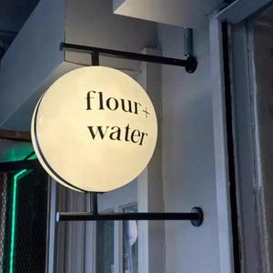 May include: A round, illuminated sign with the words "flour + water" in black lettering. The sign is mounted on a black metal frame, projecting from a light-colored wall. The sign is a shop sign.
