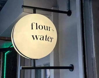 May include: A round, illuminated sign with the words "flour + water" in black lettering. The sign is mounted on a black metal frame, projecting from a light-colored wall. The sign is a shop sign.