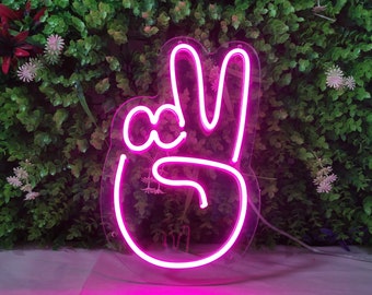 May include: Pink neon sign shaped like a hand giving the peace sign. The sign is lit up and has a clear acrylic backing.