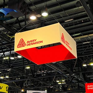 May include: A rectangular, hanging display with the Avery Dennison logo in red on a cream-colored background. The bottom interior is bright red. The display is suspended from the ceiling in a large venue.