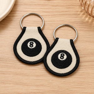 Pode incluir: Dois porta-chaves em forma de gota com uma bola de bilhar preta bordada e o número oito em branco no centro. Os porta-chaves são feitos de tecido bege com bordas pretas costuradas e incluem um anel de metal prateado.