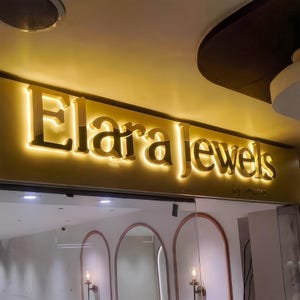 May include: Illuminated sign for "Elara Jewels" in gold lettering, above a glass storefront. The sign is lit from behind, creating a glowing effect. The words "by Seeme" are below the main text.