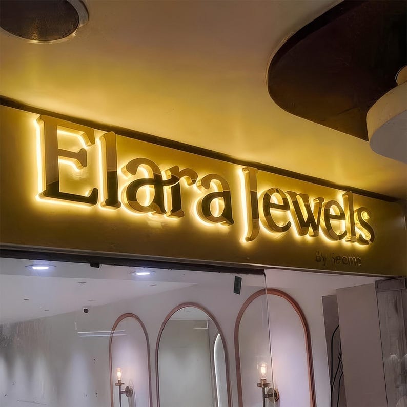 May include: Illuminated sign for "Elara Jewels" in gold lettering, above a glass storefront. The sign is lit from behind, creating a glowing effect. The words "by Seeme" are below the main text.