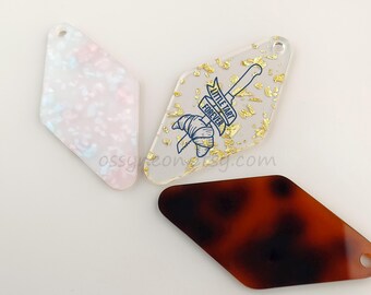 May include: Three diamond-shaped acrylic charms with different designs. One charm is clear with gold flecks and a blue graphic of a croissant and a knife with the text "Little Tart Forever". Another charm is clear with a pink and white marbled design. The third charm is brown with a tortoiseshell pattern.