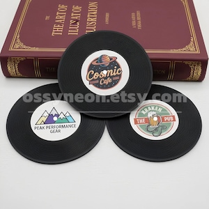 May include: Three black vinyl record-shaped coasters with white center labels. One coaster features "Cosmic Cafe" text and a planet graphic. Another has "Peak Performance Gear" text and a mountain graphic. The third has "The Kraken Pub" text and a beer graphic.