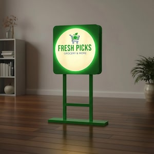 May include: A green illuminated sign displaying "FRESH PICKS GROCERY & MORE" and a shopping cart icon with an apple. The sign is square with rounded corners and is supported by a green stand. The sign is lit with a bright white light.