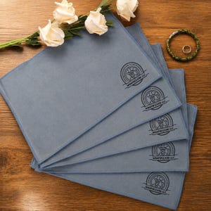 Puede incluir: Cinco paños de limpieza de microfibra azul claro con la inscripción "WRAPWIZARD LLC". Los paños están colocados sobre una superficie de madera, con una ramita de rosas blancas y una pulsera con un anillo.