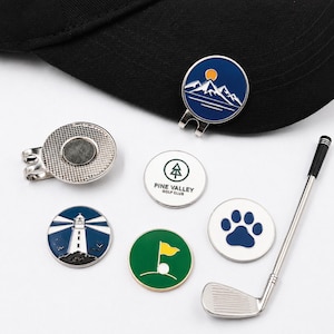 Könnte beinhalten: Eine Sammlung von Golfballmarkern mit verschiedenen Designs, darunter eine Berglandschaft, ein Leuchtturm, ein Pfotenabdruck und eine Golf-Flagge. Ein silberner Golfschläger und ein magnetischer Hutclip sind ebenfalls vorhanden. Die Marker sind in Blau-, Grün- und Weißtönen gehalten.