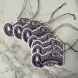 May include: A stack of six white car air fresheners with blue and red lettering that reads "Quality Body Works Est. 1978". Each air freshener has a string attached for hanging.