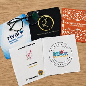 Puede incluir: Un surtido de artículos promocionales, que incluyen gafas con montura negra, paños de limpieza y una almohadilla negra con letras doradas. Los paños presentan varios diseños y textos, incluida una fecha de boda y logotipos de empresas. Los artículos están dispuestos sobre una superficie de madera.