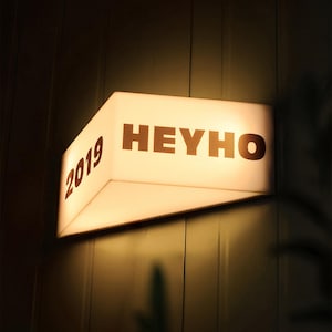 Può includere: Un'insegna rettangolare illuminata con le parole "HEYHO" e l'anno "2019" stampati in marrone. L'insegna è montata su una parete scura e strutturata ed emette una calda e invitante luce.