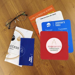 Op de afbeelding: Een assortiment kleurrijke promotieartikelen, waaronder een rode vierkant met een kerstmarktlogo, blauwe en witte placemats en een witte doek met een gouden ontwerp. Een bril ligt op een houten oppervlak.