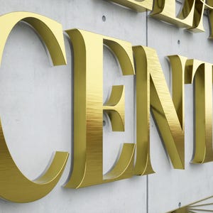 May include: Gold metallic letters spelling "CENTER" and part of another word, mounted on a light gray concrete wall. The letters have a brushed metal texture, creating a reflective surface. The overall aesthetic is modern and elegant.