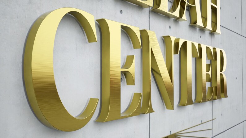 May include: Gold metallic letters spelling "CENTER" and part of another word, mounted on a light gray concrete wall. The letters have a brushed metal texture, creating a reflective surface. The overall aesthetic is modern and elegant.