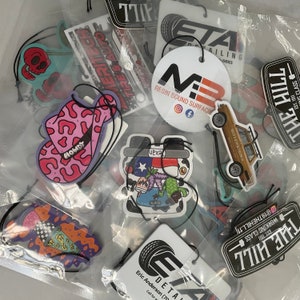 May include: Assortment of colorful car air fresheners with various designs, including a cowboy hat, a cartoon character, and a car. The air fresheners are made of paper and have a string for hanging.