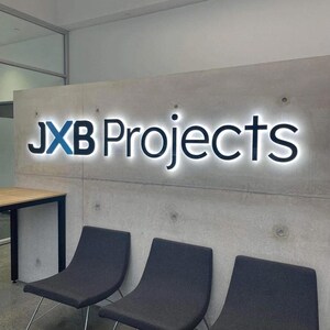 May include: The JXB Projects logo is displayed on a concrete wall with a backlit design. The logo features the text "JXB Projects" in a modern font, with the "JXB" in blue. Three dark gray chairs are in front of the wall.