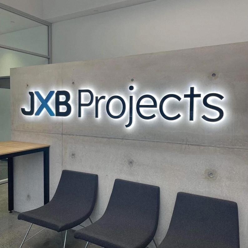 May include: The JXB Projects logo is displayed on a concrete wall with a backlit design. The logo features the text "JXB Projects" in a modern font, with the "JXB" in blue. Three dark gray chairs are in front of the wall.