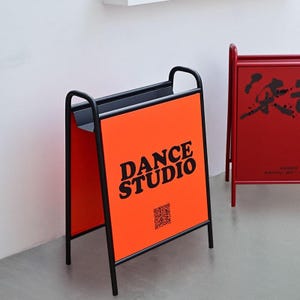 May include: An orange A-frame sign with the text "DANCE STUDIO" in black. The sign has a black metal frame and a QR code. Another red sign is visible in the background. The signs are likely for a dance studio.