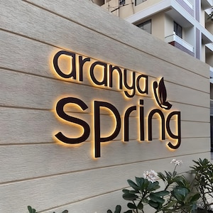 Op de afbeelding: Verlicht bord met de woorden "Aranya Spring" in een donkerbruin lettertype. Het bord is bevestigd aan een lichtbeige getextureerde muur. Een bladontwerp staat naast het woord "Spring".