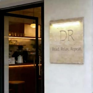 Puede incluir: Un letrero rectangular de color claro con las letras grabadas "DR" y la frase "Read. Relax. Repeat." está montado en una pared blanca. Una luz cálida ilumina el letrero desde arriba. Una puerta de cristal y el interior son visibles a la izquierda.