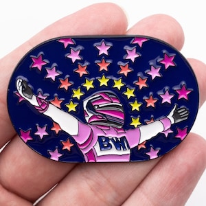 May include: A blue enamel pin with a pink and white race car driver with arms outstretched, surrounded by colorful stars. The driver is wearing a helmet and the car has the letters "BWI" on the front.