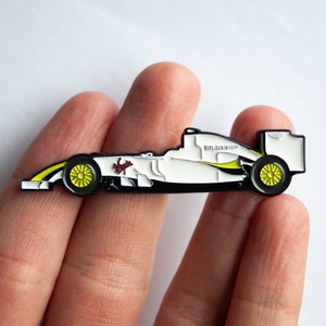 May include: A white and black enamel pin of a Formula 1 race car with yellow accents. The car has the text "BRAWN GP" on the side.