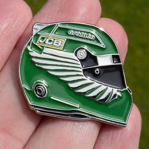 Puede incluir: Un pin de casco de carreras verde con el texto "JCB" y "aramco". El pin es un diseño de esmalte detallado, con un acabado brillante y una visera negra. El casco tiene un diseño de ala en el lateral.