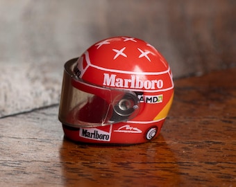 May include: A miniature red racing helmet with the "Marlboro" logo and white accents. The helmet has a clear visor and the "AMD" logo. A collectible item.