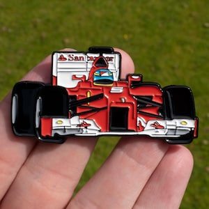 May include: A red and black enamel pin of a Formula 1 race car. The pin features the car's details, including the driver's helmet, sponsor logos, and the number 5. The pin is set against a green background.