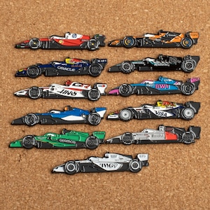 May include: A collection of Formula 1 race car pins in various colors, including red, orange, blue, green, and black. Each pin features detailed designs and sponsor logos. The pins are arranged on a corkboard surface.