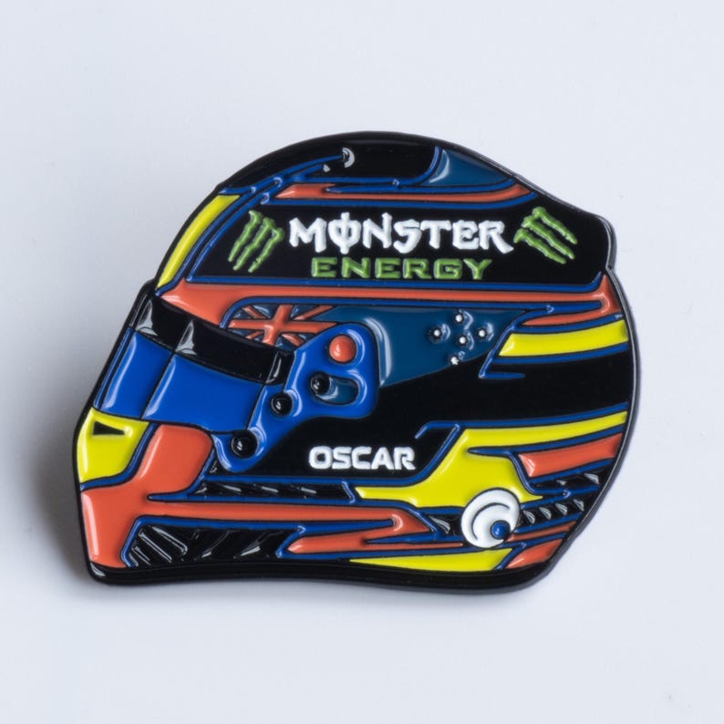 May include: A black and yellow racing helmet pin with the Monster Energy logo and the name Oscar. The helmet is blue, red, and yellow with a black background.