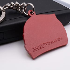 Pode incluir: Chaveiro de metal vermelho em forma de capacete de corrida com o texto "GPpins.com" gravado em relevo no lado.