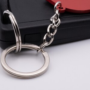 Pode incluir: Um chaveiro prateado com um charme de cora&ccedil;&atilde;o vermelho preso a um chaveiro preto.