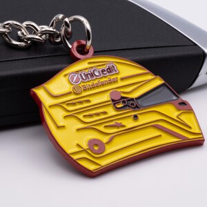 Pode incluir: Chaveiro de metal amarelo e vermelho em forma de capacete de corrida com os logotipos "UniCredit" e "Bitdefender" no lado.