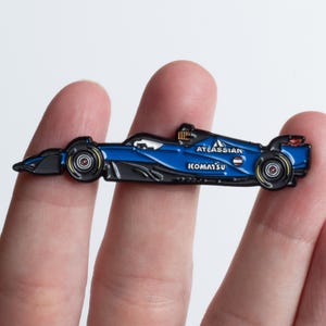 Könnte beinhalten: Ein blau-schwarzer Formel-1-Rennwagen-Pin mit dem Text "ATLASSIAN" und "KOMATSU". Der Pin wird zwischen zwei Fingern gehalten. Die Räder sind schwarz mit gelben und roten Akzenten.