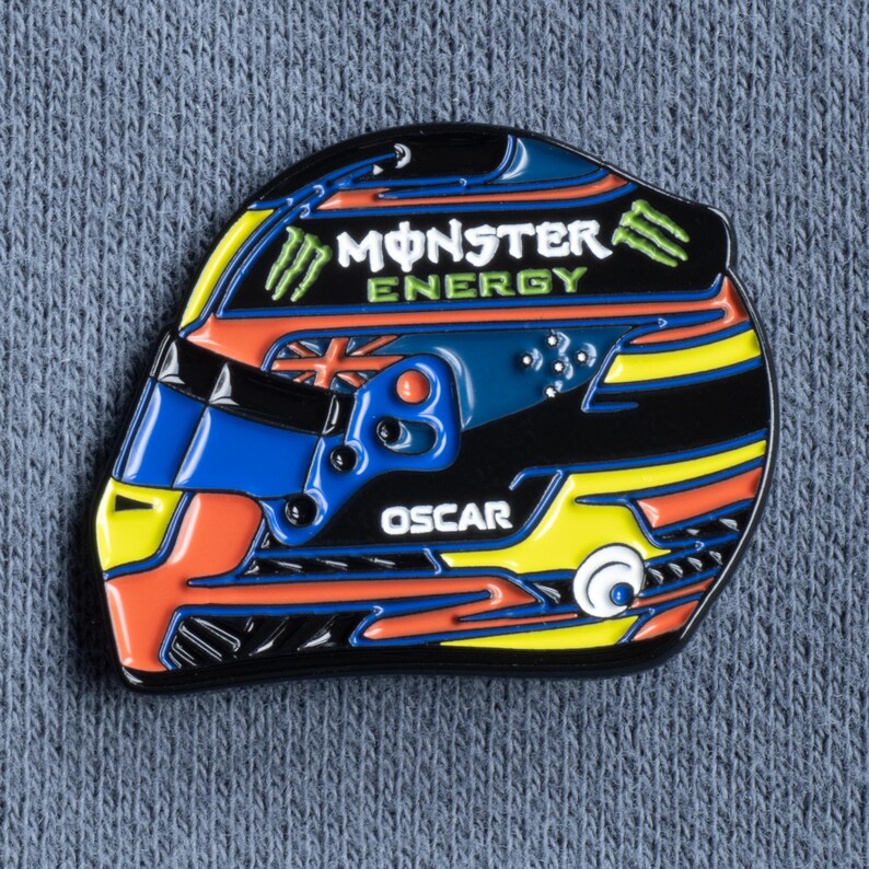 May include: A black, orange, blue, and yellow enamel pin of a racing helmet with the words "Monster Energy" and "Oscar" on it.