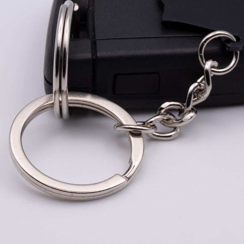 May include: Close-up of a silver key ring with a chain and clasp, attached to a black car key fob. The key ring is circular and has a shiny, metallic finish. The chain is made of small, interconnected links.
