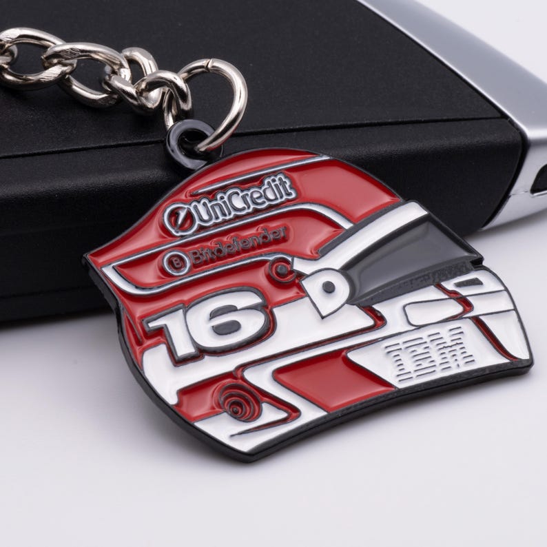 May include: A red, white, and black helmet-shaped keychain with the number 16 and the UniCredit logo. The keychain is made of metal and has a silver chain and clasp.
