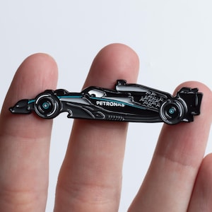 Peut inclure: Épingle de voiture de course de Formule 1 noire et sarcelle avec le mot "PETRONAS" sur le côté. L'épingle est tenue entre deux doigts sur fond blanc. La voiture a des roues détaillées et des caractéristiques aérodynamiques.