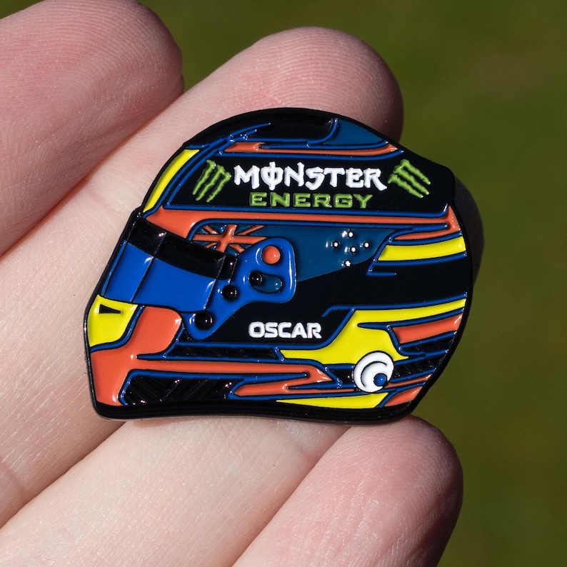 May include: A black, blue, yellow, and orange enamel pin of a racing helmet with the words "Monster Energy" and "Oscar" on it.