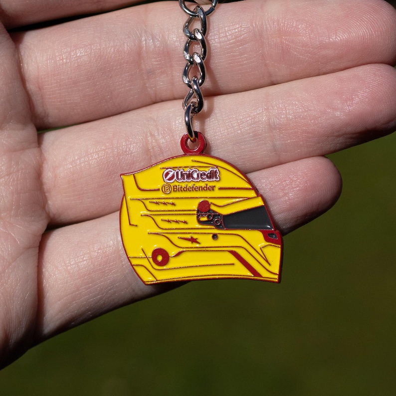 Pode incluir: Um chaveiro de metal amarelo e vermelho em forma de capacete de corrida. O capacete tem o texto "UniCredit" e "Bitdefender".