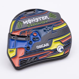 May include: A black and yellow enamel pin shaped like a racing helmet. The helmet has a Monster Energy logo, the text "OSCAR", and a blue, red, and yellow design.
