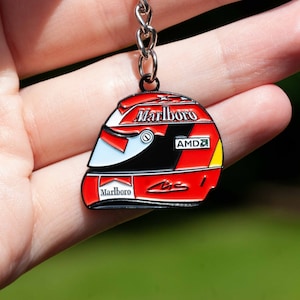 Könnte beinhalten: Ein roter, schwarzer und weißer Schlüsselanhänger aus Metall mit einem Rennhelm mit den Worten "Marlboro" und "AMD" darauf.