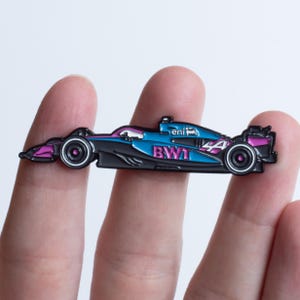 Może przedstawiać: Przypinka z bolidem Formuły 1 w kolorze niebieskim, czarnym i różowym. Przypinka zawiera numer 44 i logo BWT. Samochód ma elegancki design ze szczegółowymi kołami i elementami aerodynamicznymi. Przypinka jest trzymana między dwoma palcami.