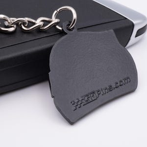 May include: A dark gray keychain with a silver chain and ring. The keychain has a textured surface and the text "GPPins.com" is visible. The keychain is resting on a black surface.