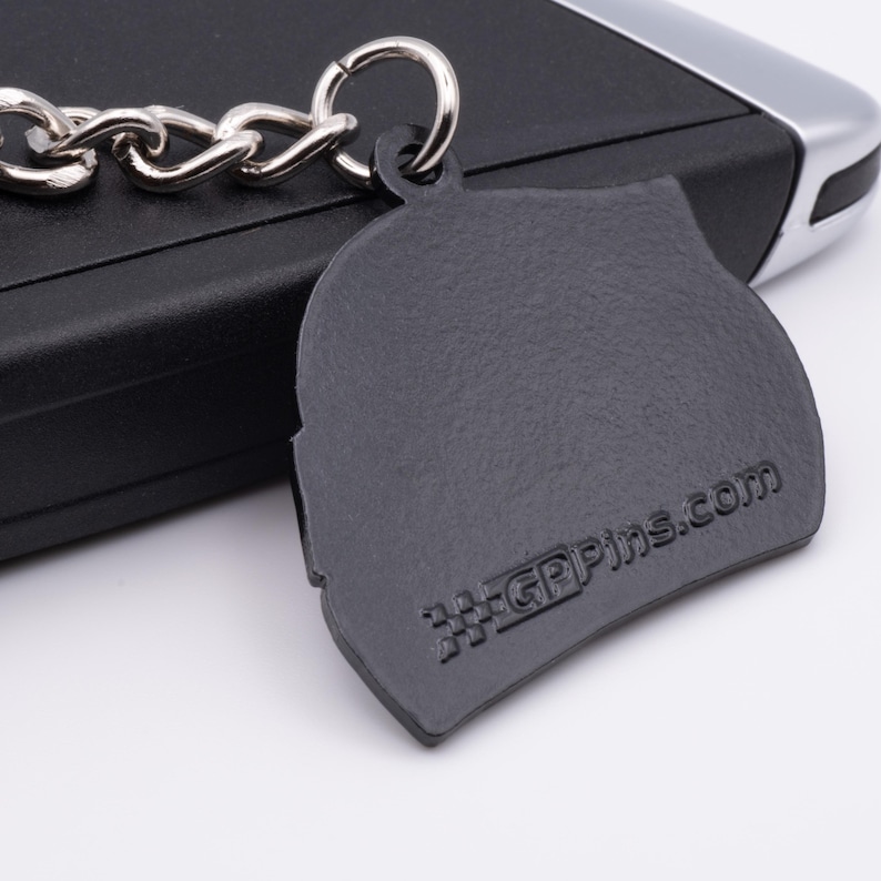 May include: A dark gray keychain with a silver chain and ring. The keychain has a textured surface and the text "GPPins.com" is visible. The keychain is resting on a black surface.