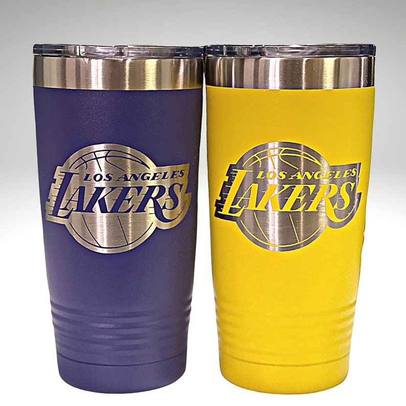 May include: Two insulated tumblers, one purple and one yellow, featuring the Los Angeles Lakers logo. The logo is a silver-colored emblem with the team name and a basketball design. Both tumblers have a silver rim and lid.