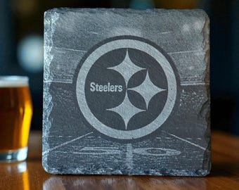 May include: Square slate coaster with the Pittsburgh Steelers logo, a circular design with the word "Steelers" in white. The coaster has a rough, textured edge. The background is blurred.
