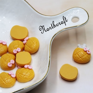 May include: A white ceramic dish with a gold rim contains nine yellow and orange resin food charms with pink bows. The dish has a handle with the word "Northcraft" written on it.